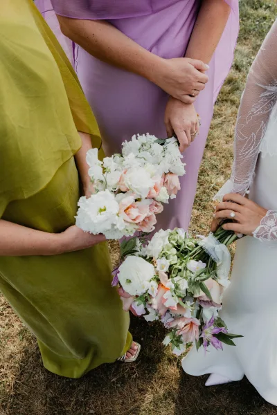 Bridesmaid bouquets of white and blush flowers with greenery, ribbon wraps, and wedding rings shown in hand on a grass lawn