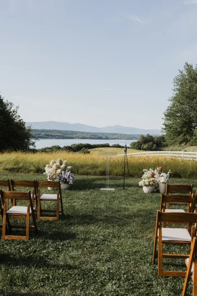 Outdoor ceremony setup with ceremony aisle chairs, pastel florals in urn planters, and a clear acrylic podium on a lawn by lake and mountains