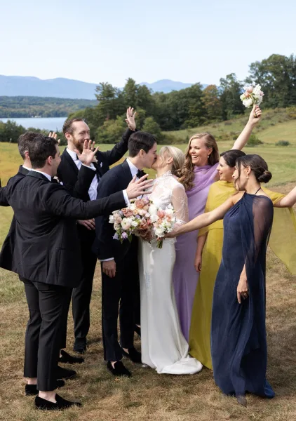 Wedding party portrait with bride and groom kissing as bridesmaids and groomsmen cheer in a grassy meadow with mountains and a lake behind them