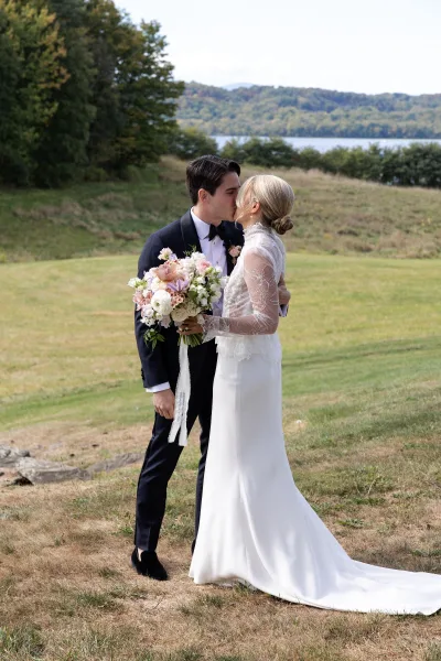 Wedding kiss portrait of bride and groom kissing, her lace-sleeved dress and bouquet in hand, with lake, hills, and trees behind