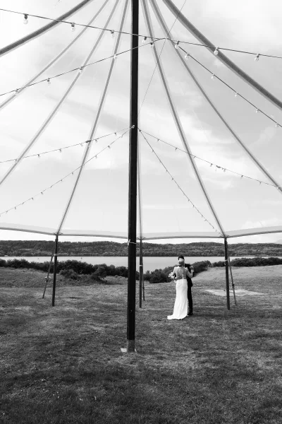 First dance outdoors as bride in wedding dress and groom in suit sway beneath string lights under a tent by a lakeside lawn with hills