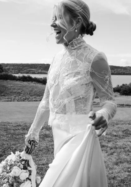 Bridal portrait of a laughing bride holding a bouquet, wearing a high-neck lace dress with drop earrings in a grassy lakeside field under clouds