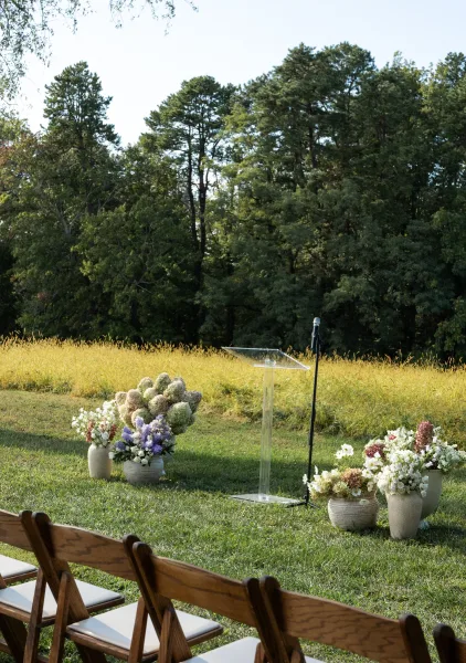 Ceremony setup for an outdoor wedding ceremony with wood folding chairs and white cushions, clear acrylic podium and flowers on a meadow lawn near trees