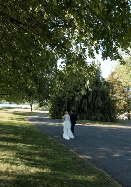 Couple walking away, newlyweds walking together under a tree canopy on a park road, bride in veil with bouquet beside groom in suit