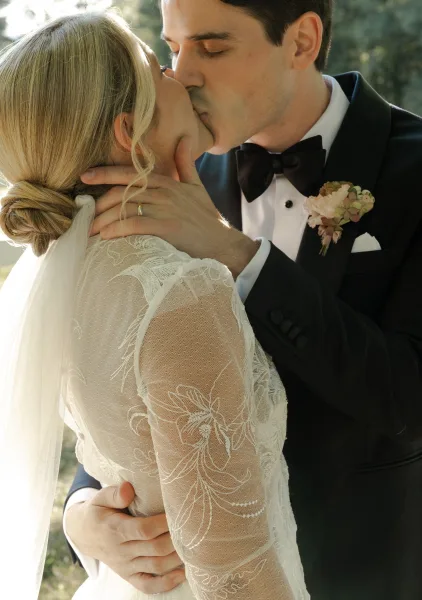 Wedding kiss as groom holds bride’s face, her lace long-sleeve dress and veil visible, black-tie tuxedo against sunlit greenery backdrop