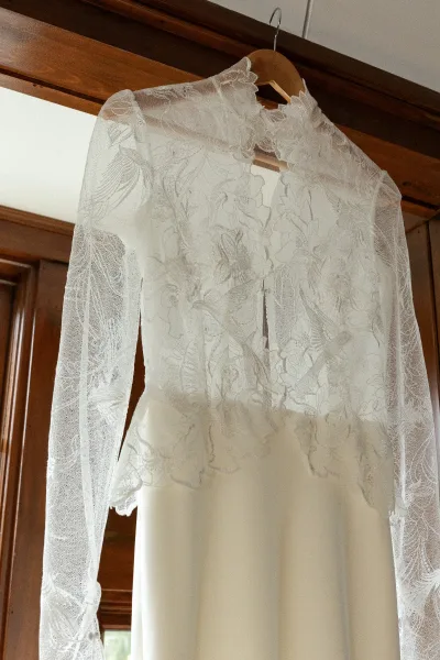 Wedding dress with long sleeves, floral lace applique and sheer neckline on a wooden hanger by a sunlit window and door frame