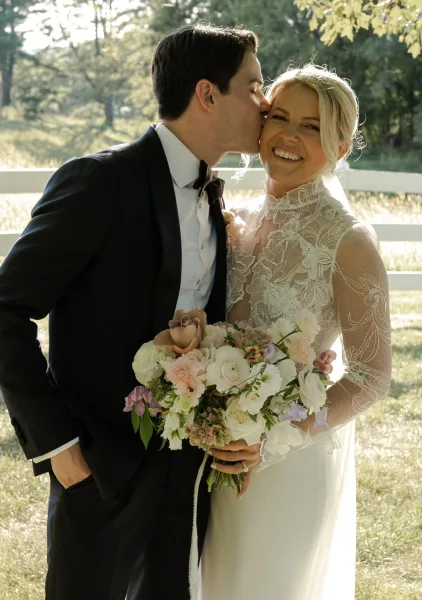 Wedding couple portrait with groom kissing bride’s cheek as she smiles, holding a blush bouquet in golden sunlight by a white fence