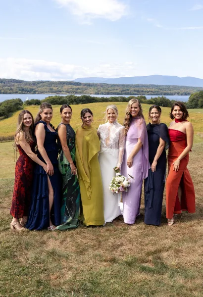 Bridesmaids portrait with bride and colorful mismatched dresses, holding a white bouquet in a grassy field by a mountain lake under blue sky
