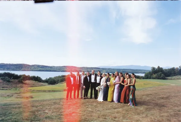 Wedding party photo of bride and groom with wedding party in a lineup on a grassy hillside by a lake and mountains, bridesmaids with bouquets
