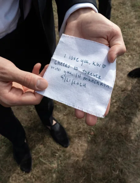 Groom gift note on a white fabric square with handwritten wedding note beside a suit jacket and dress shirt cuff on a grass lawn