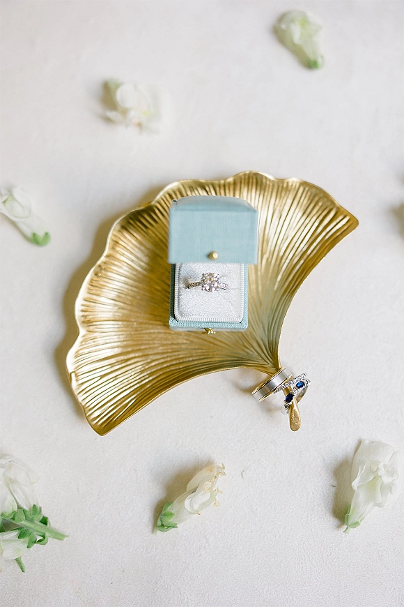 Wedding rings and engagement ring flat lay in a blue velvet ring box on a gold leaf dish with white flowers over soft white fabric backdrop