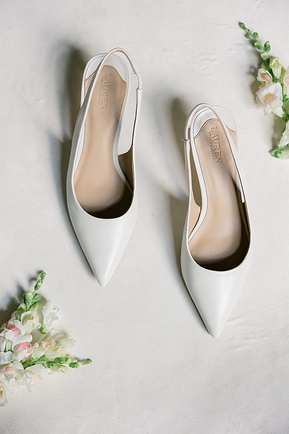 Bridal shoes in a flatlay of white wedding heels with pointed toes and slingback straps, styled with floral sprigs on light fabric
