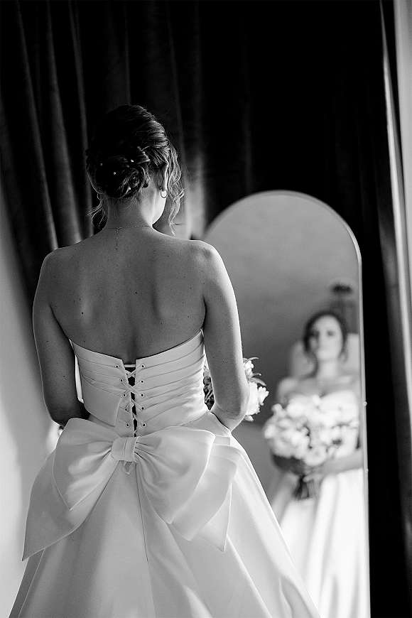 Bridal portrait of a bride in mirror holding bouquet, showing strapless corset-back dress with oversized bow in window-lit dressing room