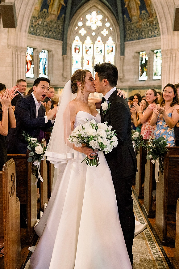 Wedding kiss as the bride and groom share a just married kiss in a church aisle, veil flowing amid stained glass and cheering guests