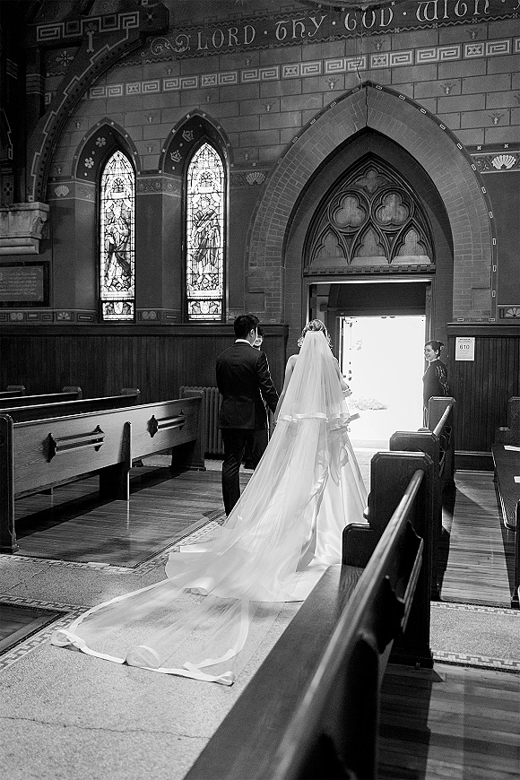 Processional moment as bride walking down aisle from behind, cathedral veil trailing past pews in a stained-glass church interior