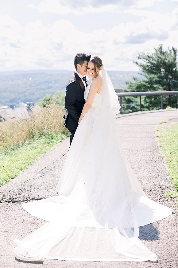 Couple portrait of bride and groom embrace as he gives a forehead kiss, her cathedral veil draped over shoulders on a mountain overlook path