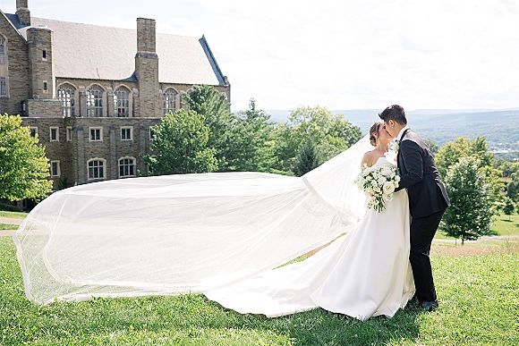 Wedding kiss portrait of bride and groom kissing, her long veil trailing behind, holding white rose bouquet on lawn by stone building