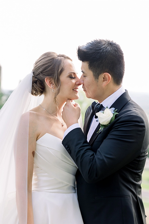 Wedding kiss portrait close up of bride and groom nose kiss, groom hand on her chin, veil and tuxedo against bright greenery sky
