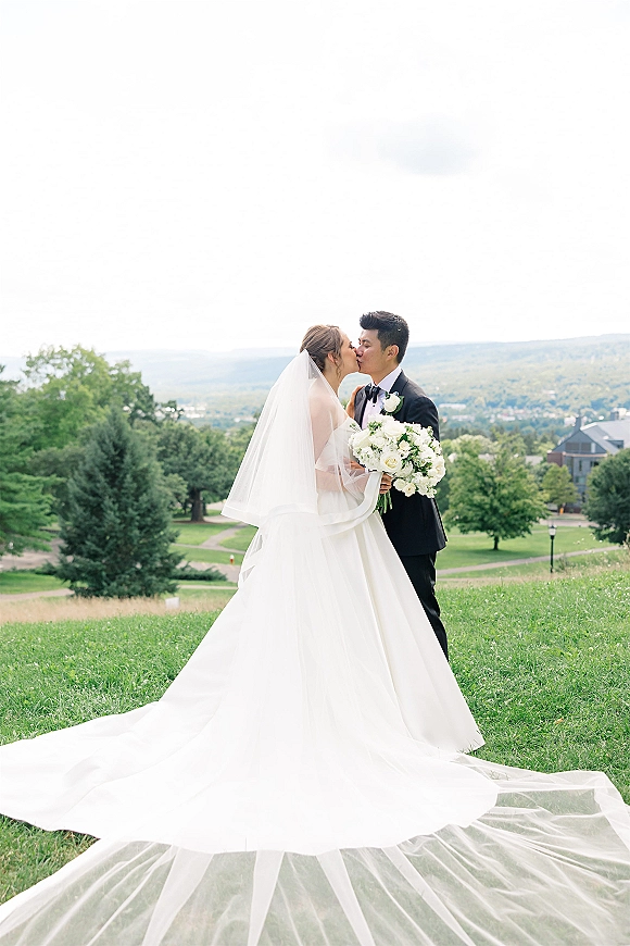Wedding kiss portrait of bride and groom kissing, her veil and white rose bouquet visible on a grassy hill with trees and sky behind