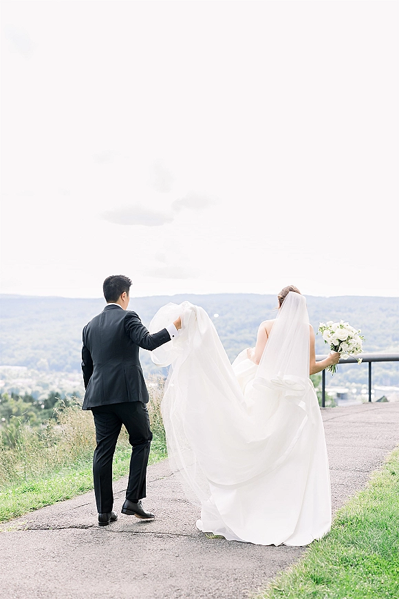 Couple portrait of bride and groom walking away, groom lifting her dress train, long veil flowing on a mountain overlook path.