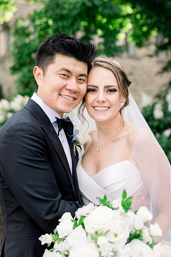 Couple portrait of bride and groom hugging, her veil draped over shoulders as they smile in a garden with trees behind