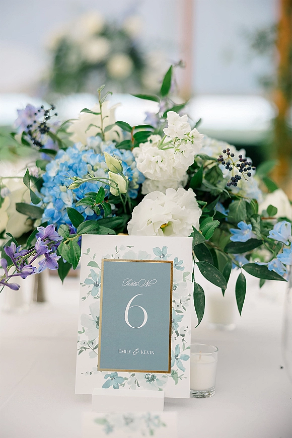 Wedding table number in a gold frame with blue hydrangeas and white blooms on a white tablecloth, set in a softly blurred reception tent.