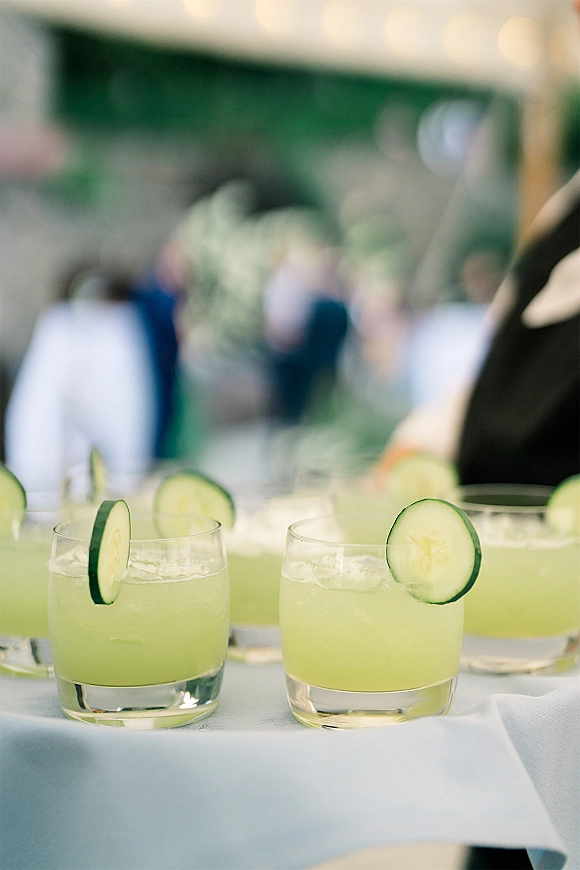 Signature cocktails in lowball glasses with cucumber slices and ice on a light blue tablecloth at an outdoor reception with greenery