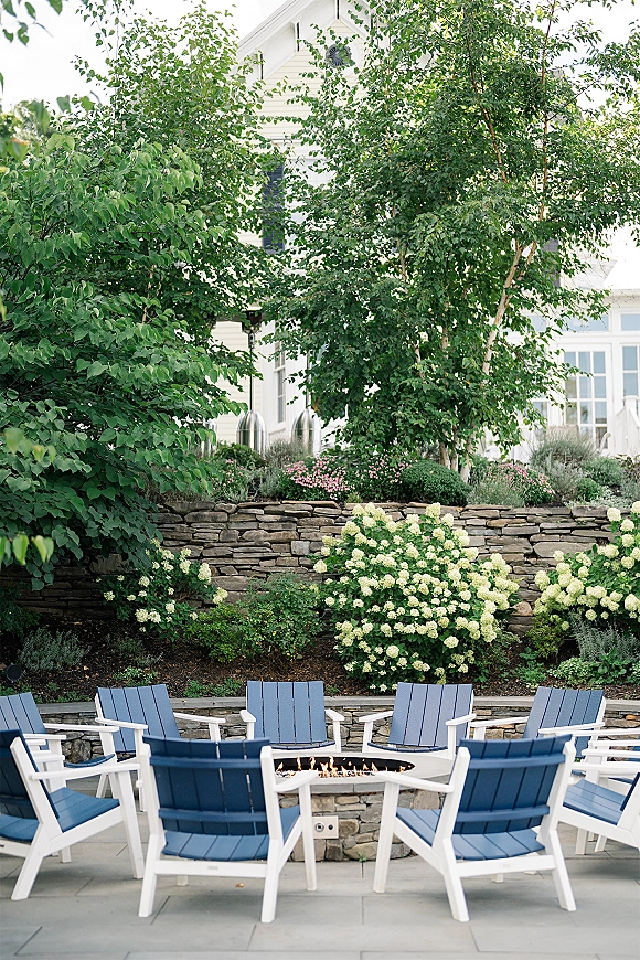 Outdoor lounge area with wedding cocktail hour lounge seating, Adirondack chairs around a fire pit on a stone patio by hydrangeas
