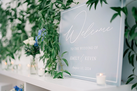Wedding welcome sign on clear acrylic with calligraphy lettering, greenery garland, blue and white flowers, and votive candles on a shelf ledge