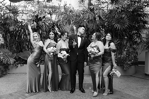 Wedding party portrait of groom with bridesmaids, holding bouquets in high-slit satin dresses, posing playfully in a tropical greenhouse backdrop