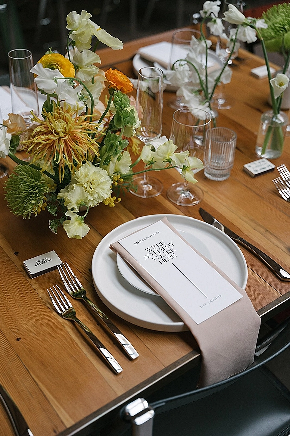 Reception tablescape on a wood wedding table setting with white plates, blush napkins, menu cards, champagne flutes, and floral centerpiece on dark chairs
