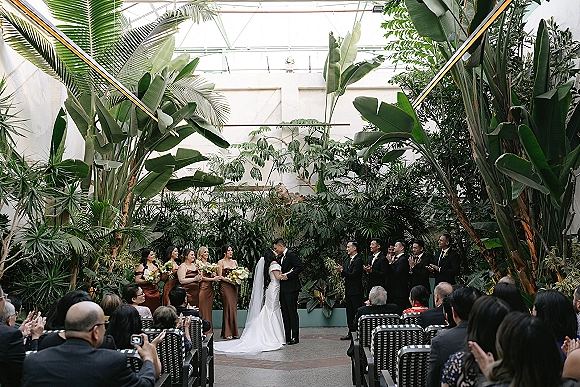 Ceremony kiss as bride in veil and groom in black tuxedo share a kiss at the altar under a glass greenhouse ceiling amid tropical plants