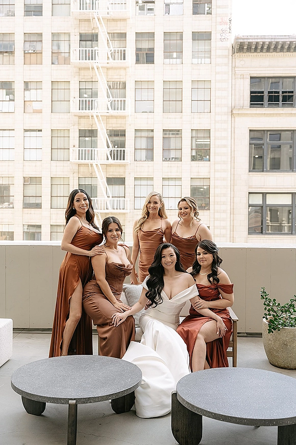 Bridesmaid group photo of the bride with bridesmaids in copper satin dresses seated on a terrace lounge, city windows behind