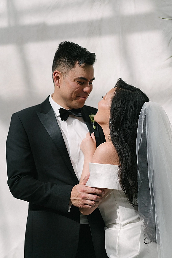 Couple portrait of bride and groom in a newlywed embrace, her veil draped over an off-shoulder satin dress against a white wall