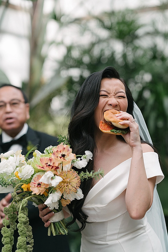 Bride portrait of her eating a burger while holding a bouquet, veil and off-the-shoulder dress in a tropical greenhouse setting
