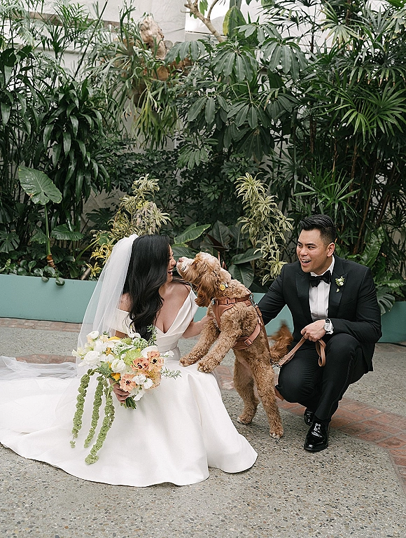 Wedding couple with dog posing as the groom holds the leash and the bride in an off-shoulder dress holds her bouquet in a lush courtyard patio