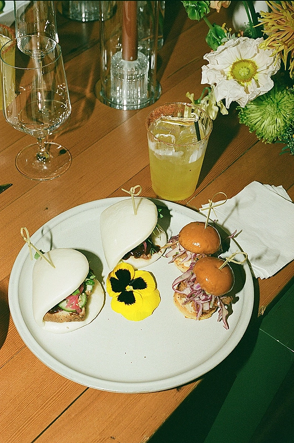 Wedding cocktail hour food and wedding passed appetizers on a white plate with bao buns, mini sliders, and a cocktail with ice on a wooden table