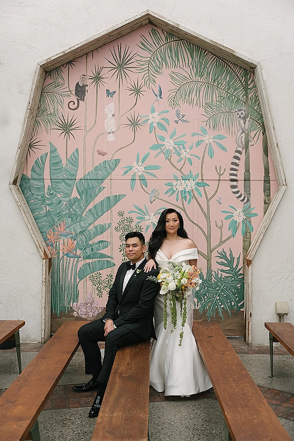 Couple portrait of bride in an off-the-shoulder gown holding a bouquet beside groom in black tux seated on a bench by a tropical mural wall