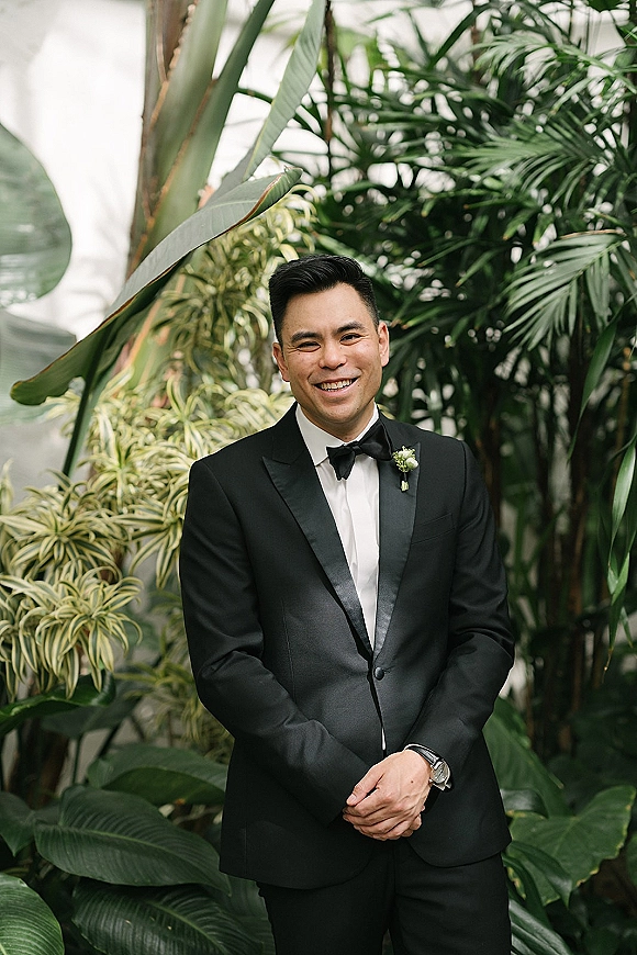 Groom portrait in a black tuxedo with bow tie and boutonniere, hands clasped and wristwatch visible amid tropical greenery