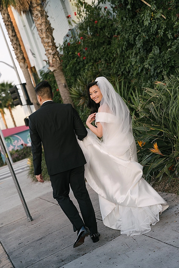 Couple portrait of bride and groom walking away on a city sidewalk, bride looking back with veil, holding dress train near palm trees