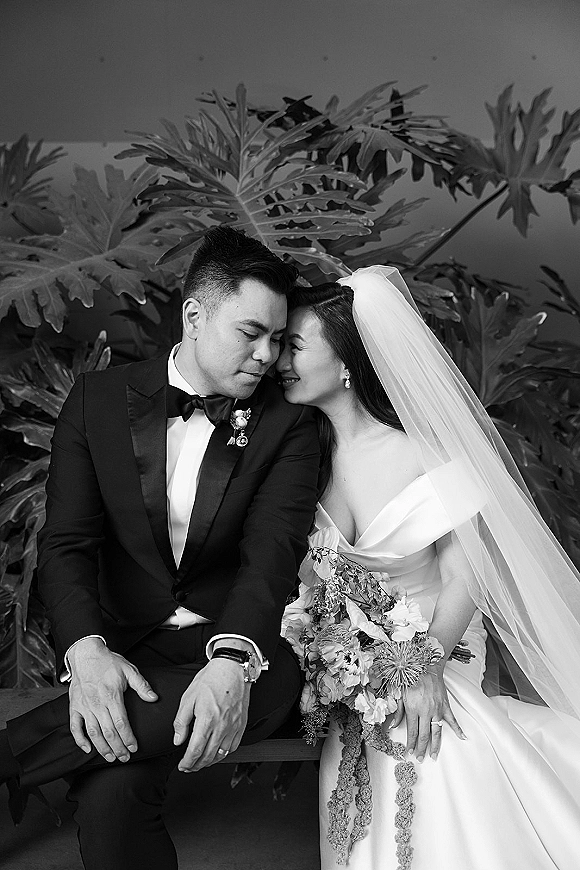 Couple portrait in a black and white wedding portrait, bride and groom touching foreheads on a bench before tropical leaves, veil draped