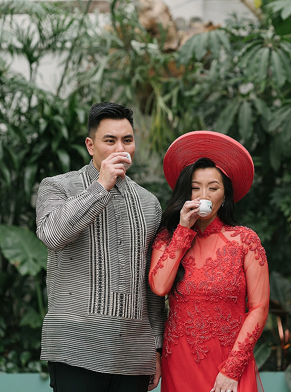 Couple portrait of bride and groom drinking coffee, her red embroidered ao dai and wide brim hat, toasting with espresso in a tropical greenhouse