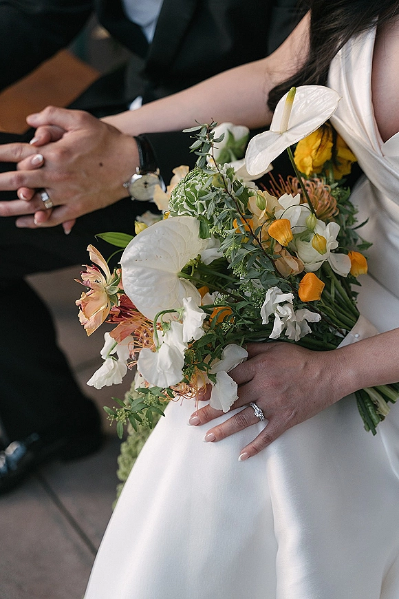 Bridal bouquet with anthurium wedding bouquet blooms in orange and white, held by bride and groom with rings near ceremony chairs