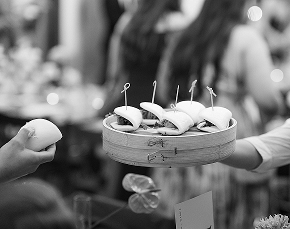 Wedding appetizers displayed as bao buns in a bamboo steamer with cocktail picks, with reception guests and blurred tables behind