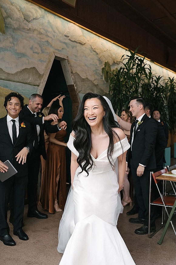 Bride portrait of a laughing bride in an off the shoulder wedding dress and long veil, walking past cheering guests by a mural wall indoors