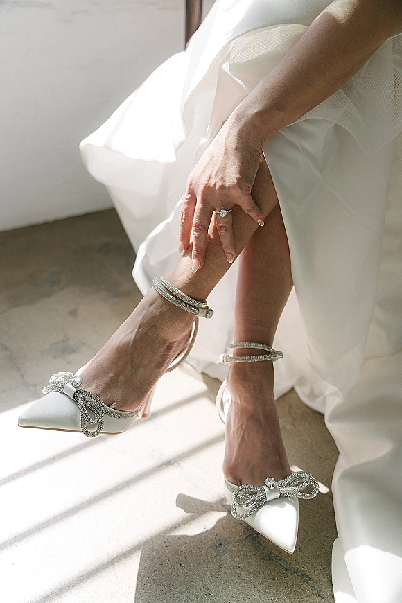 Bridal shoes, white wedding heels with rhinestone bow and ankle strap as the bride adjusts them in soft window light on a concrete floor