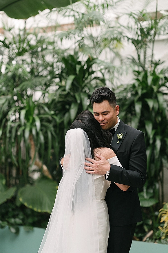 Couple portrait of bride and groom embracing, her long veil draped over a strapless dress as he smiles amid tropical greenery backdrop