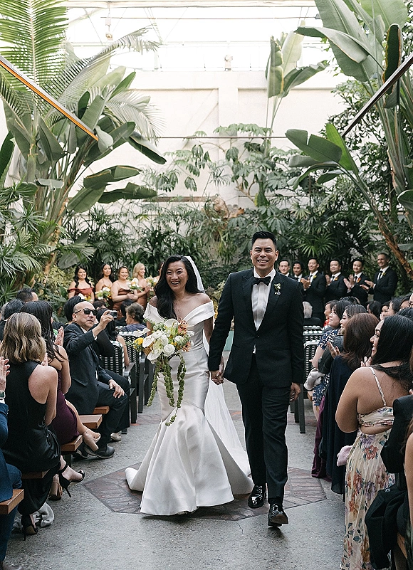 Wedding recessional as bride and groom walking aisle hand in hand, bouquet and cathedral veil flowing in a tropical greenhouse with glass ceiling