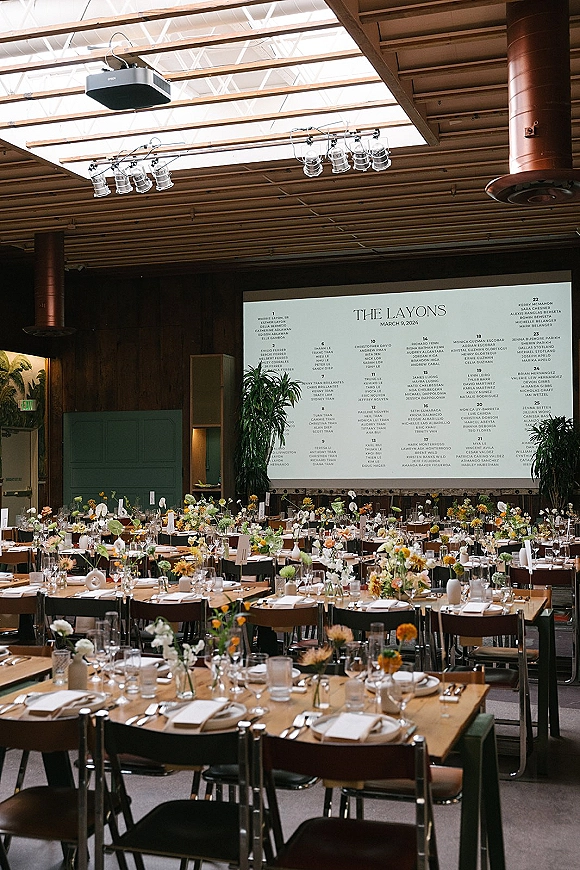 Reception tablescape with wedding reception table decor on wood banquet tables, bud vase florals, taper candles, and place cards under skylit beams