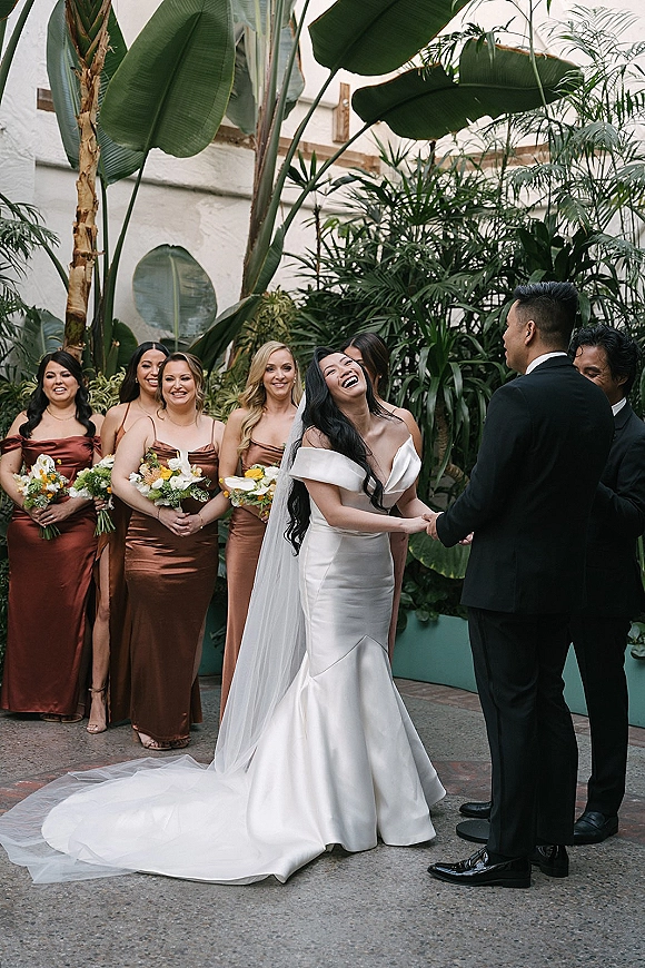 Ceremony moment as bride laughs holding hands with groom at the altar, cathedral veil and bridesmaids with bouquets in a tropical greenhouse